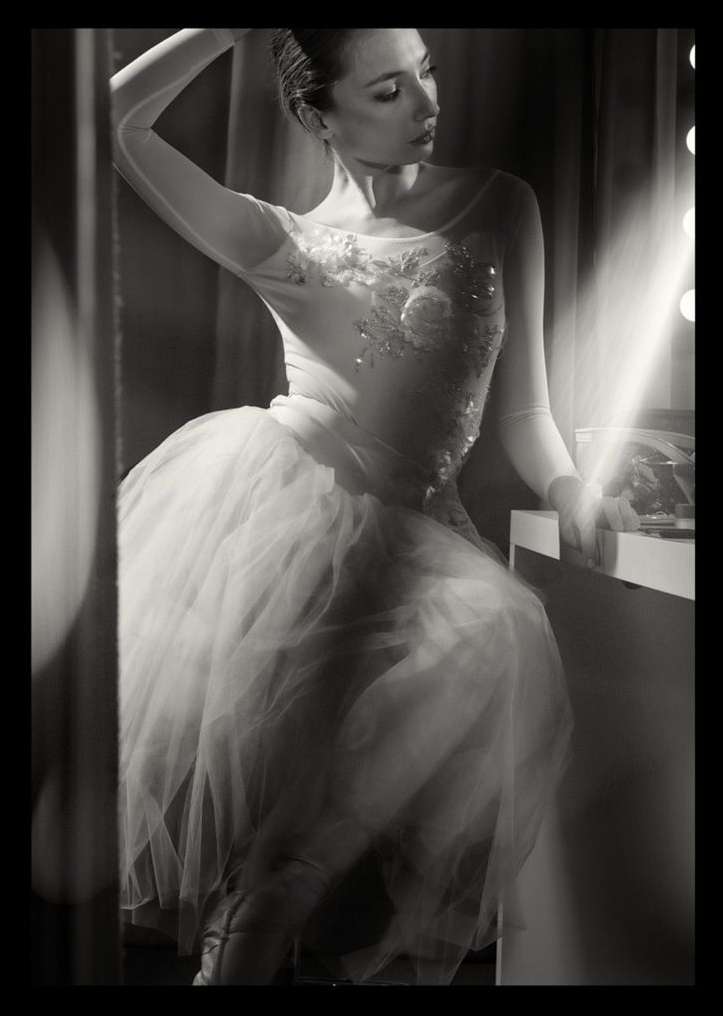 Black and white portrait of a young adult woman in a white tutu and bodice, sitting at a vanity mirror with stage lighting.