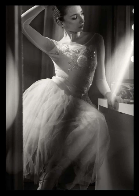 Black and white portrait of a young adult woman in a white tutu and bodice, sitting at a vanity mirror with stage lighting.