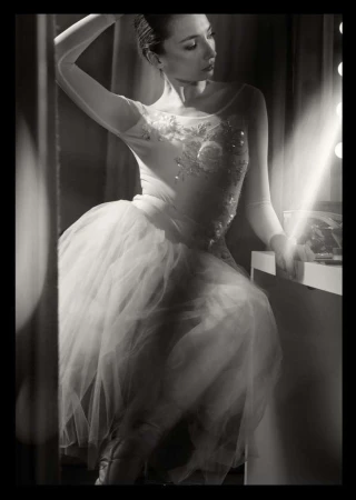 Black and white portrait of a young adult woman in a white tutu and bodice, sitting at a vanity mirror with stage lighting.