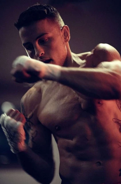A bare-chested adult male with tattoos, clenched fists, and hand wraps is shown in a medium indoor shot with a dark background.