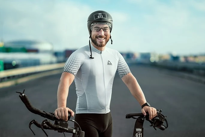 Smiling adult man in cycling gear and helmet poses with his bicycle on an empty road with buildings blurred in the background.