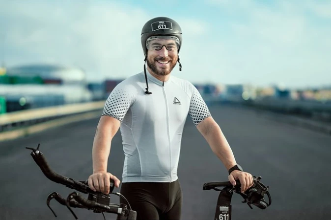 Smiling adult man in cycling gear and helmet poses with his bicycle on an empty road with buildings blurred in the background.