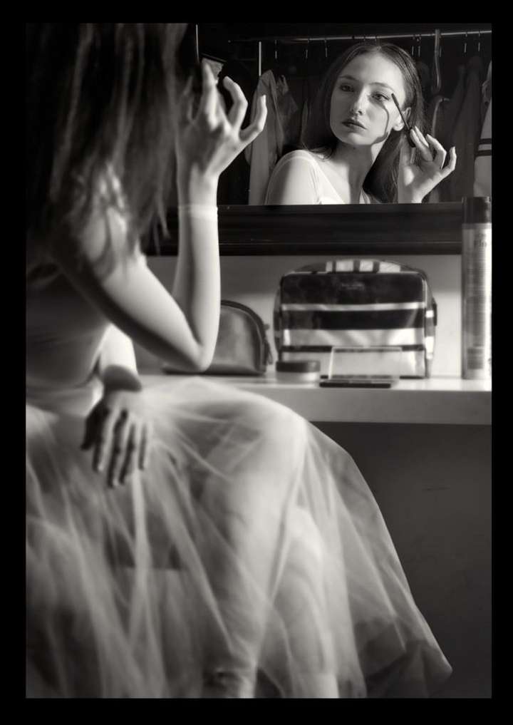 Monochrome portrait of a young woman applying makeup in front of a mirror, reflecting her face and the beauty products on the table.
