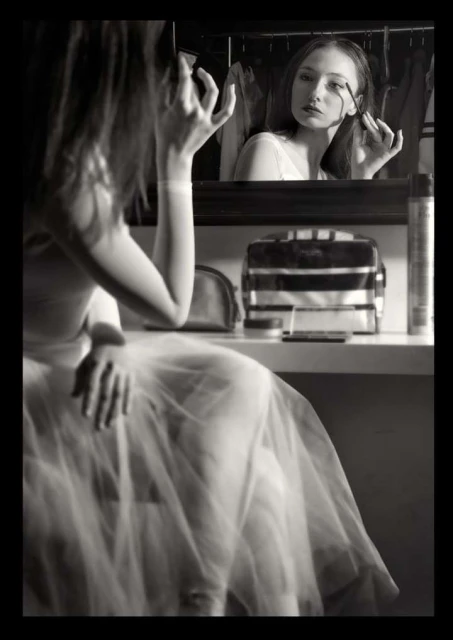 Monochrome portrait of a young woman applying makeup in front of a mirror, reflecting her face and the beauty products on the table.