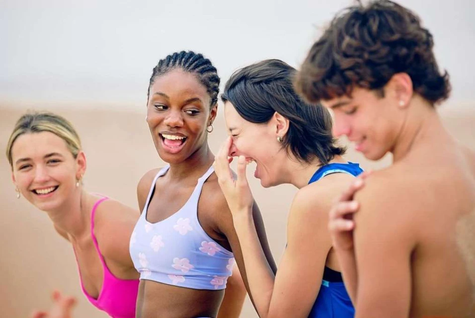 A group of four young adults, two women and one man, laughing and smiling outdoors on a sandy beach with light-colored clothing.