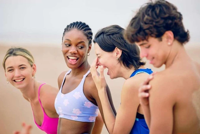 A group of four young adults, two women and one man, laughing and smiling outdoors on a sandy beach with light-colored clothing.