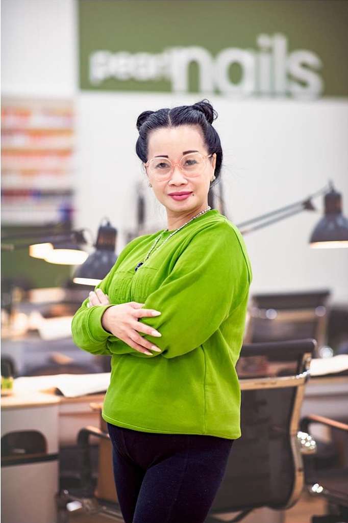 Woman in a green sweater stands with arms crossed in a nail salon, showcasing a professional and welcoming atmosphere.