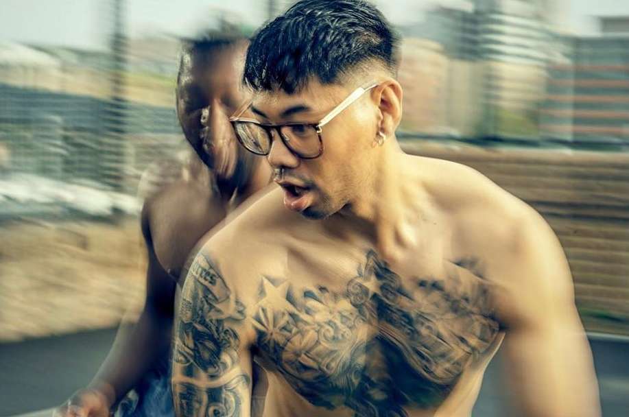 Tattooed man with glasses, shirtless, close-up shot. Another person blurred in the background. Urban setting. Daylight.