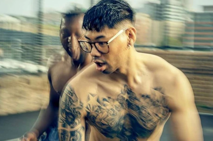 Tattooed man with glasses, shirtless, close-up shot. Another person blurred in the background. Urban setting. Daylight.