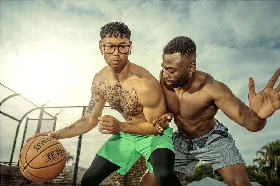Two shirtless men playing basketball outdoors. One man dribbles the ball, the other defends. Bright daylight and partly cloudy sky.
