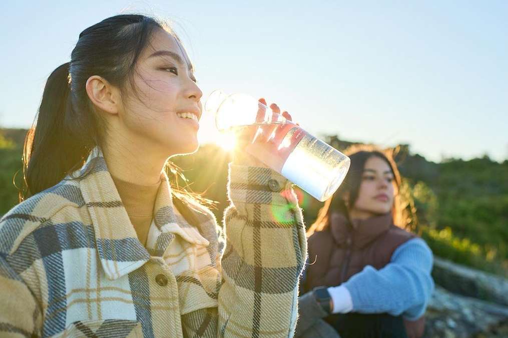 A young woman drinks from a clear plastic water bottle in the sunshine, with another woman seated in the background.