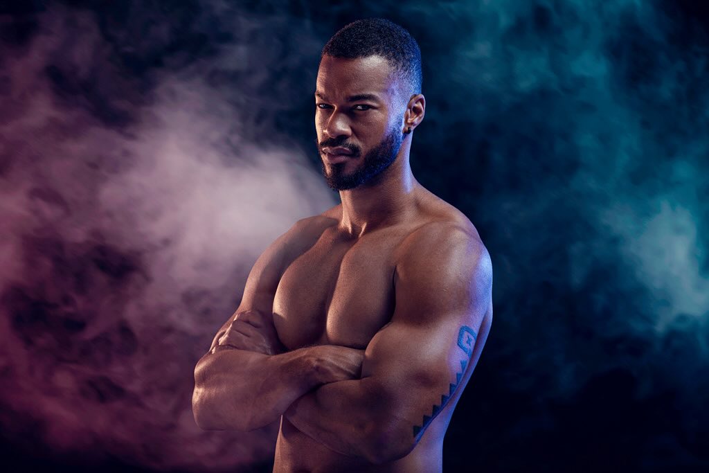 Shirtless African American man with muscular build poses with arms crossed against a backdrop of purple and teal smoke in studio lighting.