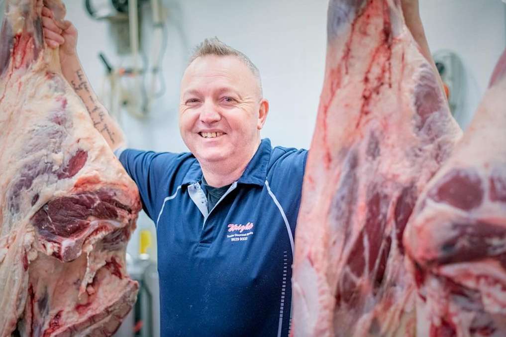 Smiling butcher in a blue polo shirt stands with sides of raw meat hanging in a white-walled shop, creating a vivid and striking visual.