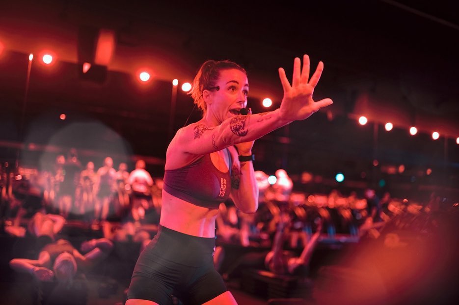Fitness instructor leading a group cycling class in a dark, red-lit studio, motivating participants. High-energy workout scene.