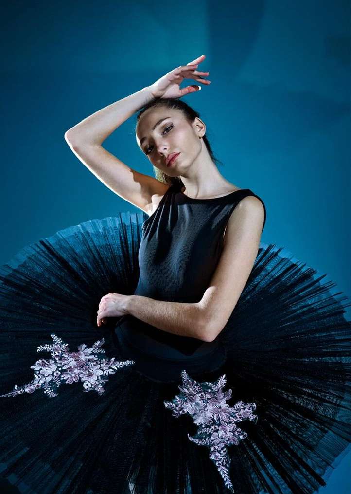 Young ballerina poses gracefully in a black tutu with floral appliques against a dark teal backdrop in a studio-lit shot.