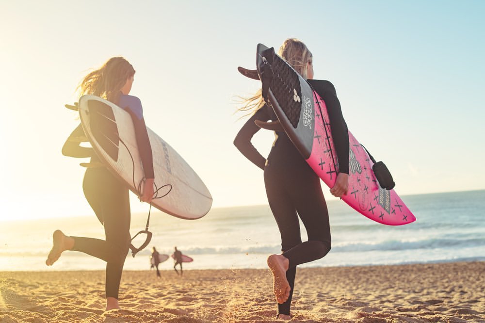 Two female surfers in wetsuits walking on a beach at sunset, carrying surfboards, with the ocean and other surfers in the background.