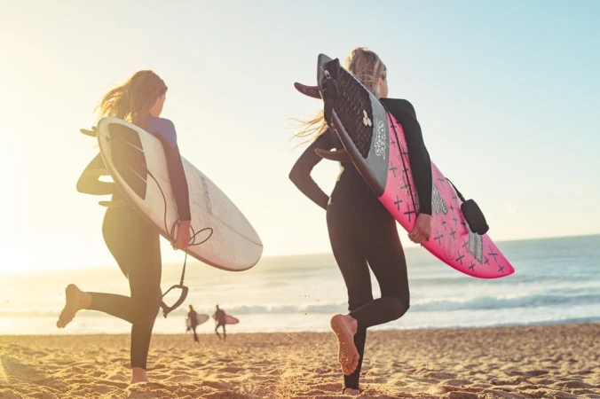 Two female surfers in wetsuits walking on a beach at sunset, carrying surfboards, with the ocean and other surfers in the background.