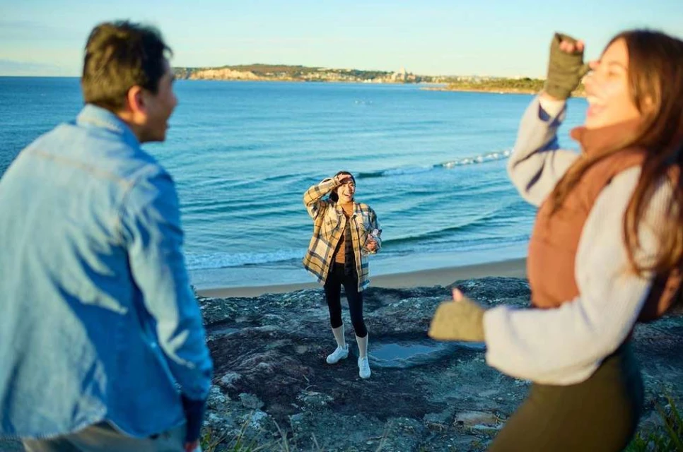 Three people standing on a rocky cliff overlooking a scenic coastline with ocean views, blue sky, and distant land.
