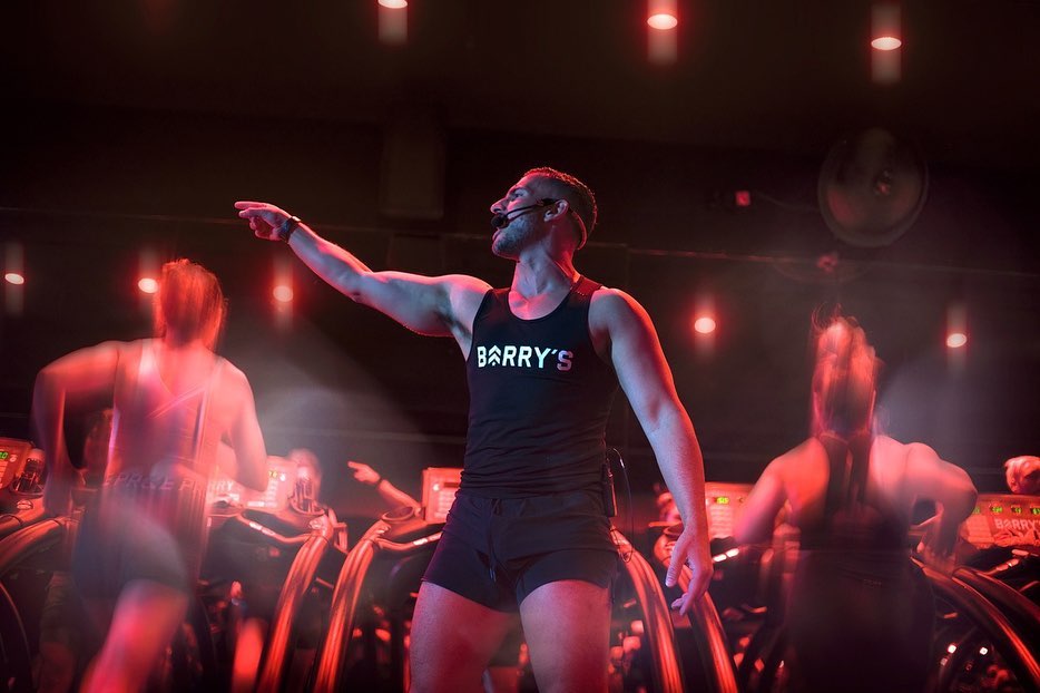 Fitness instructor leads class on treadmills in a dark studio, illuminated by red lighting, wearing Barry's branded athleticwear.