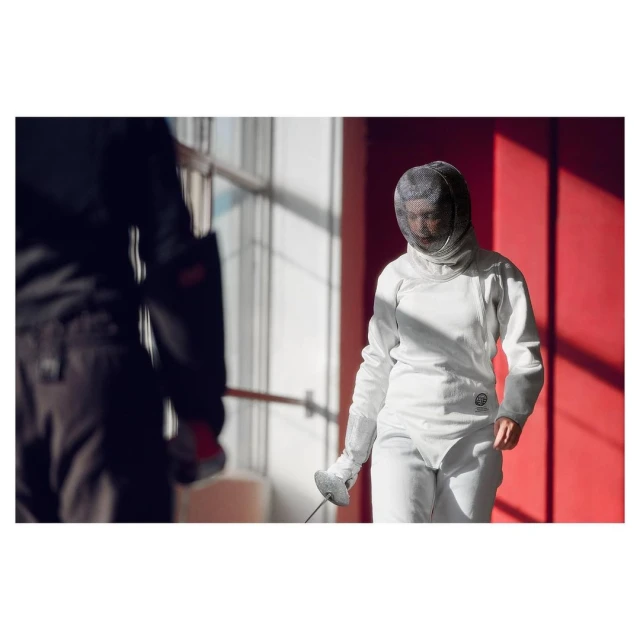 Woman fencer in white uniform and mask holds foil in a brightly lit indoor space, another person is visible in the blurred background.