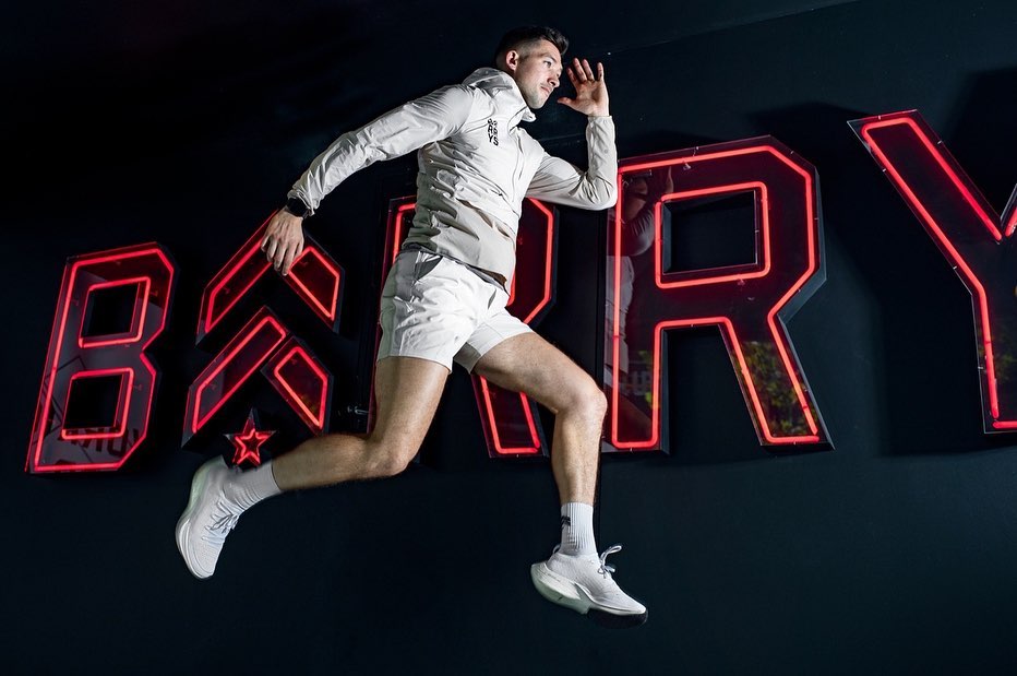 A fit man jumps energetically in a studio setting, wearing athletic wear and sneakers, with vibrant red neon sign in the background.