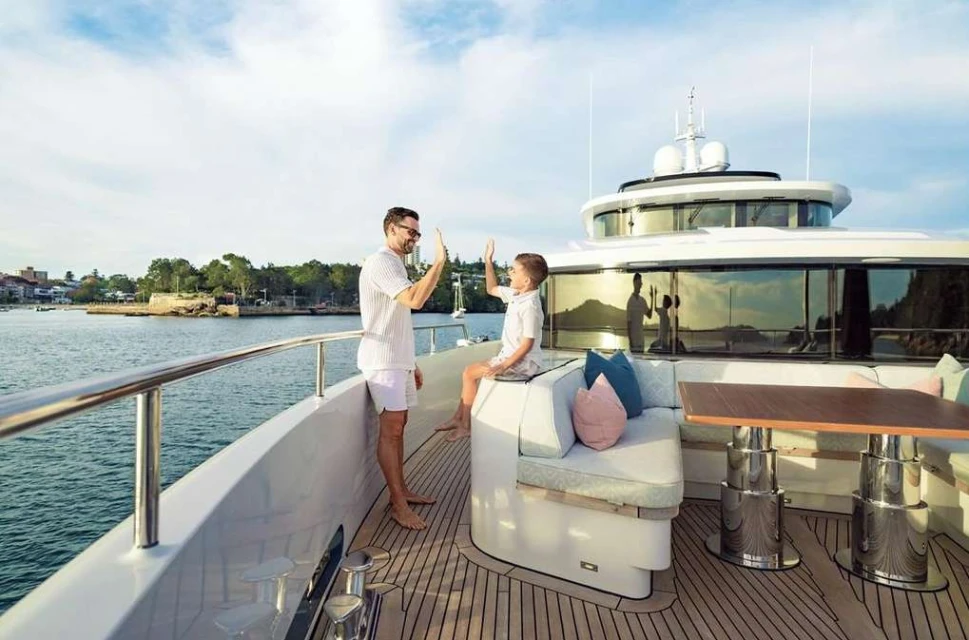 A father and son on a yacht give each other a high five, with coastal scenery visible in the background under a bright blue sky.