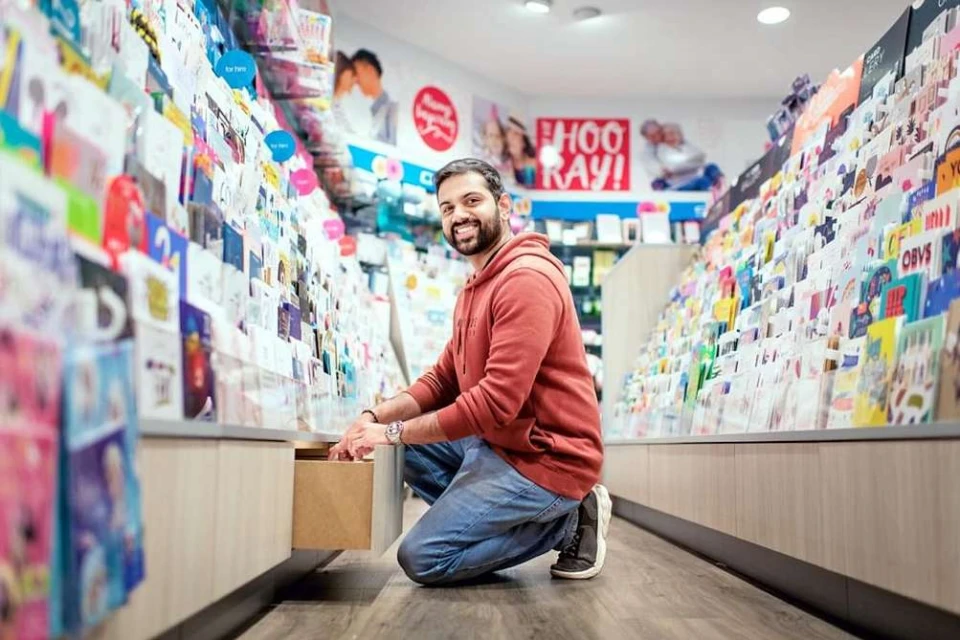Man crouching in a shop, smiling at the camera while opening a display drawer filled with greeting cards and stationery.
