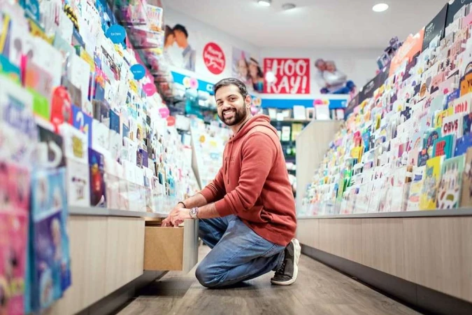 Man crouching in a shop, smiling at the camera while opening a display drawer filled with greeting cards and stationery.