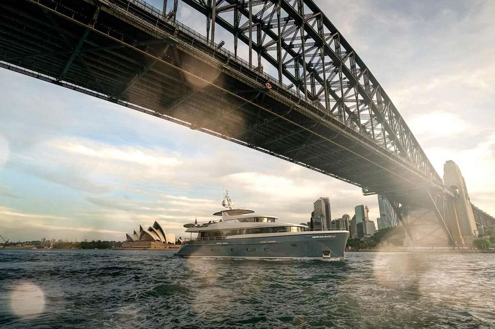 Luxury yacht sails beneath Sydney Harbour Bridge, with the Sydney Opera House and city skyline in the background on a sunny day.