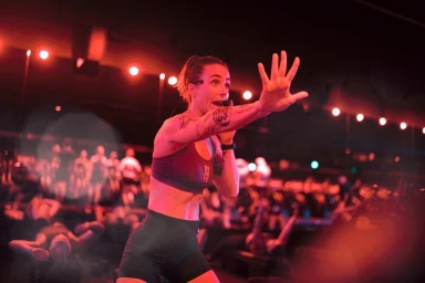 Fitness instructor leading a class in a red-lit studio, holding hand up with open palm while wearing activewear and a headset microphone.