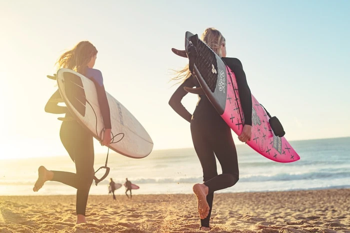 Two women in wetsuits walking on a sandy beach carrying surfboards toward the ocean during sunset