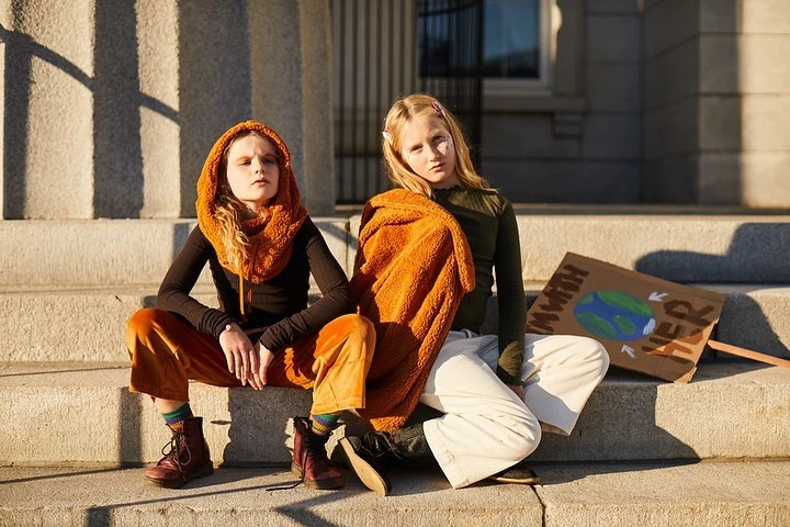 Two young girls sit on steps in vintage-inspired clothing, beside a sign with a painted Earth, in front of a stone building.