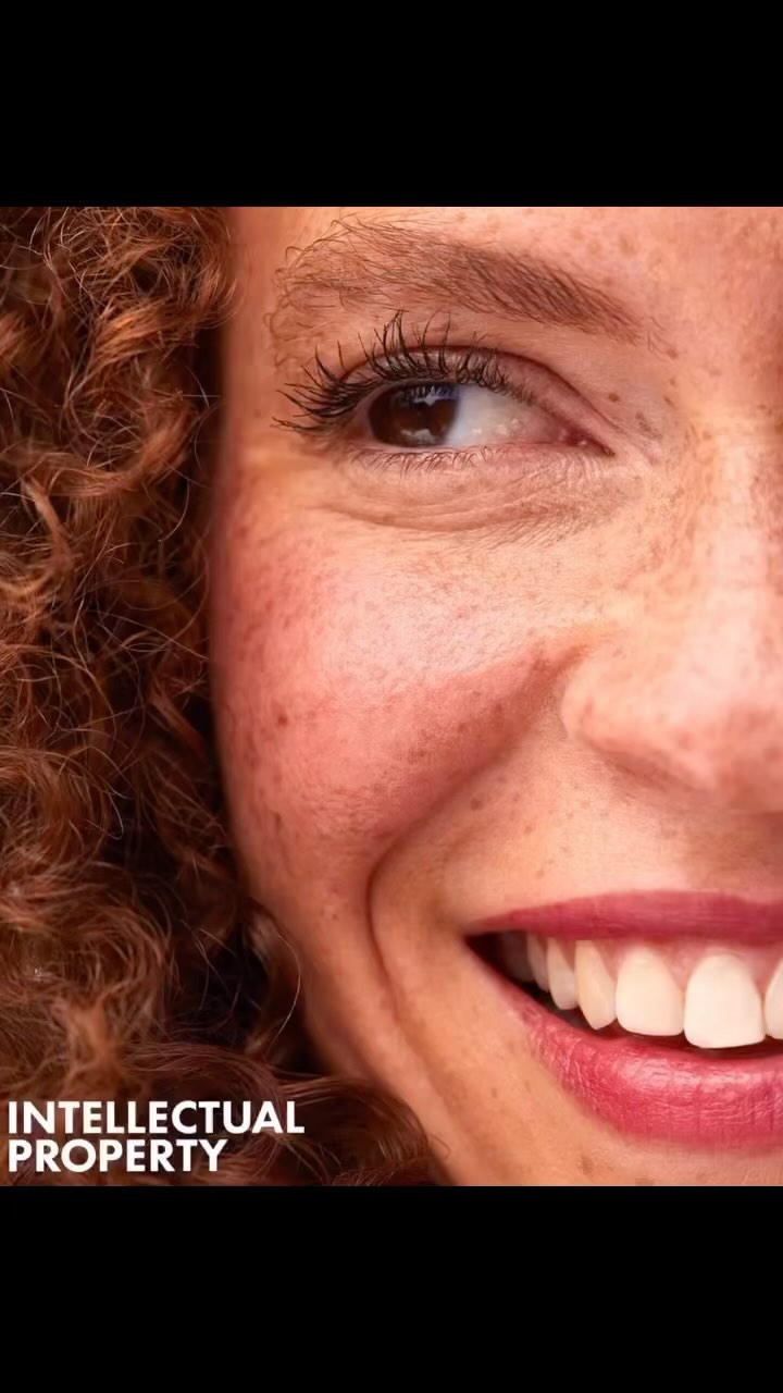 Close-up portrait of a smiling adult female with curly red hair, freckles, and pink lipstick. The image captures a portion of her face.