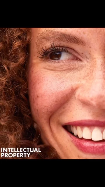 Close-up portrait of a smiling adult female with curly red hair, freckles, and pink lipstick. The image captures a portion of her face.