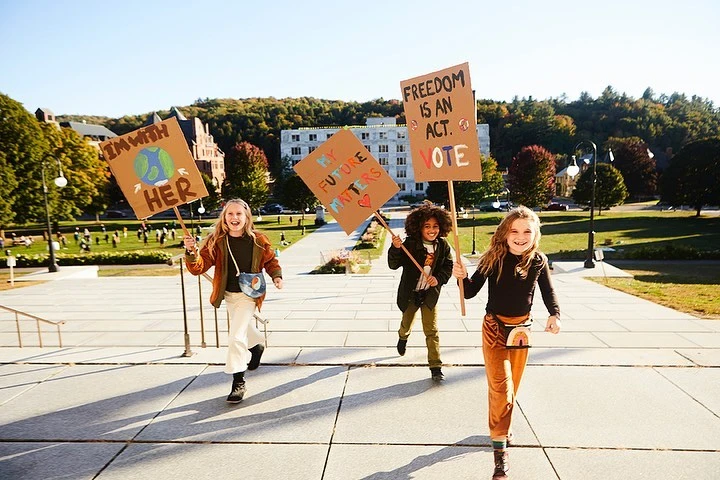 Three smiling girls run with handmade signs advocating for the Earth and voting, in front of a stately building and lush greenery.