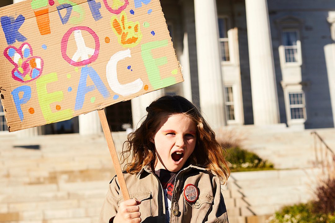 Child holds a colorful 'Kind to Peace' sign during a protest outside a building with large columns, advocating for peace and kindness.
