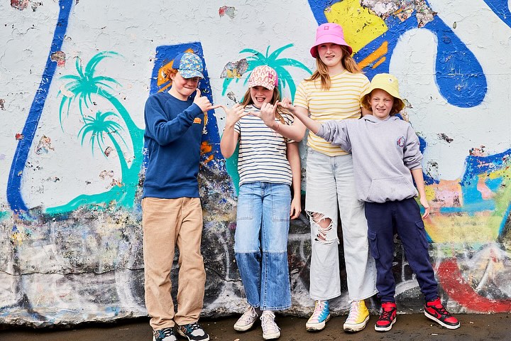 Group of four young friends stand in front of graffiti wall wearing bright hats, striped shirts, jeans and sneakers in outdoor setting.