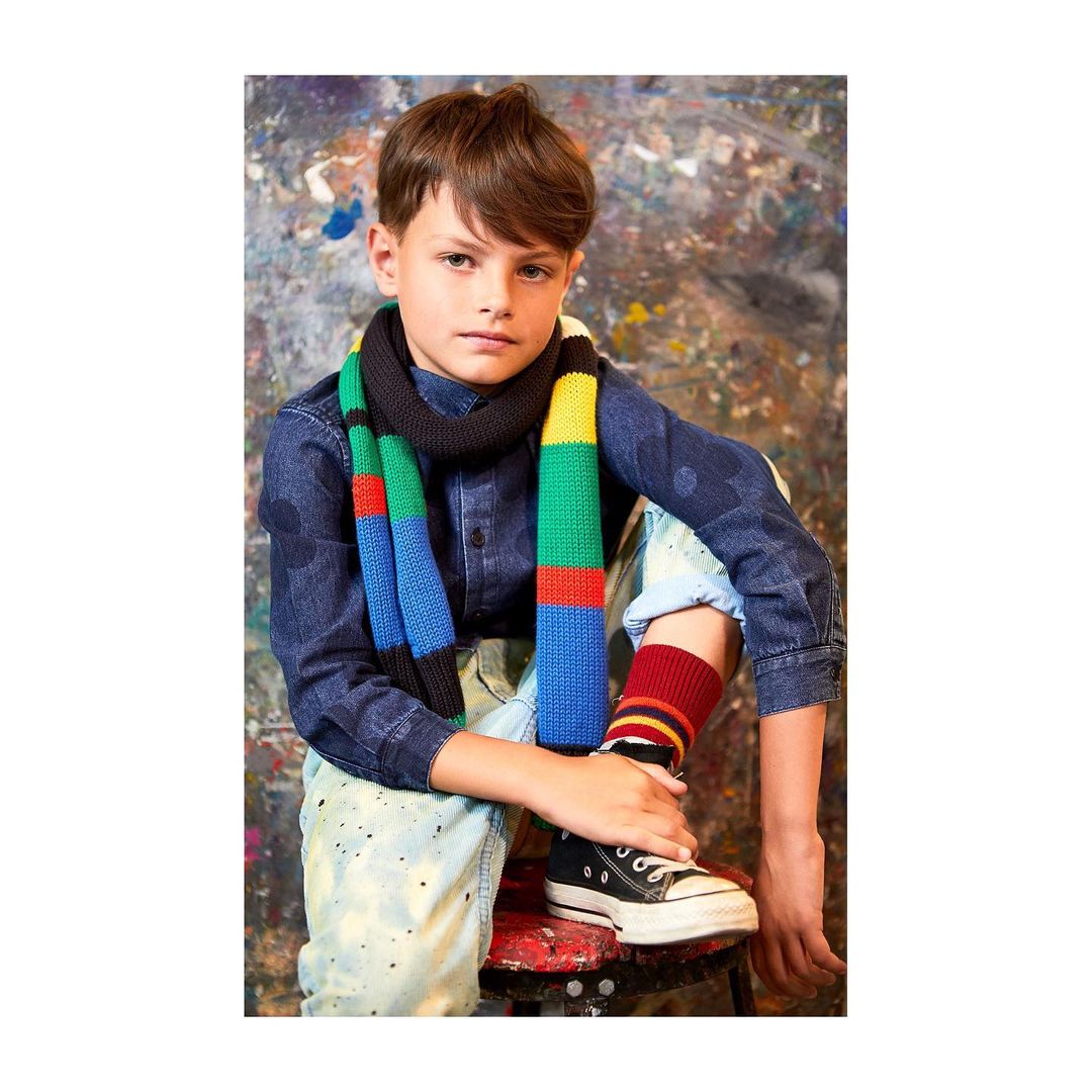 Boy seated indoors against painted backdrop, wearing a blue shirt, colorful scarf, socks, and black sneakers, looking at the camera.