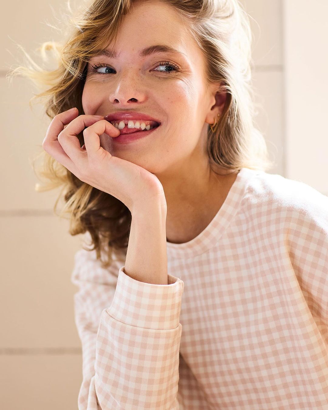 Close-up of a smiling woman with blonde hair wearing a pink and white gingham shirt, with her hand near her face.