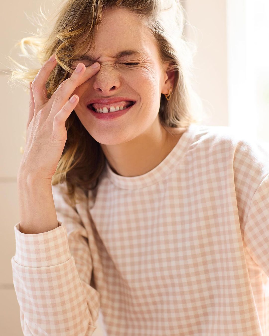Smiling adult female with hand near face, posing in a photo studio wearing a beige and white gingham shirt, indoor setting.