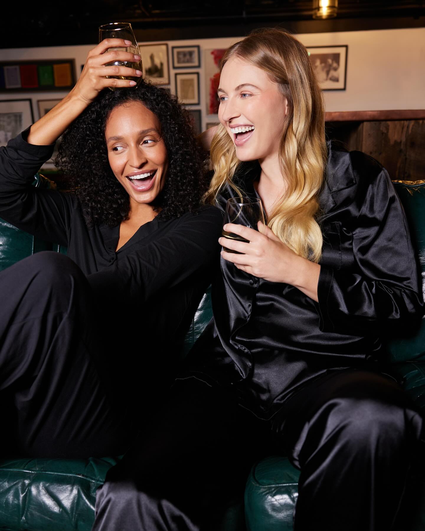 Two women laugh together on a green couch indoors. One woman has dark curly hair, and the other has long blond hair. Both wear black pajama sets.