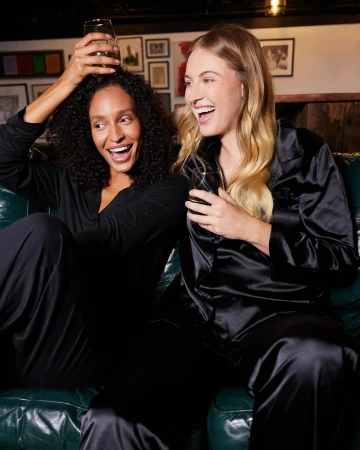 Two women laugh together on a green couch indoors. One woman has dark curly hair, and the other has long blond hair. Both wear black pajama sets.