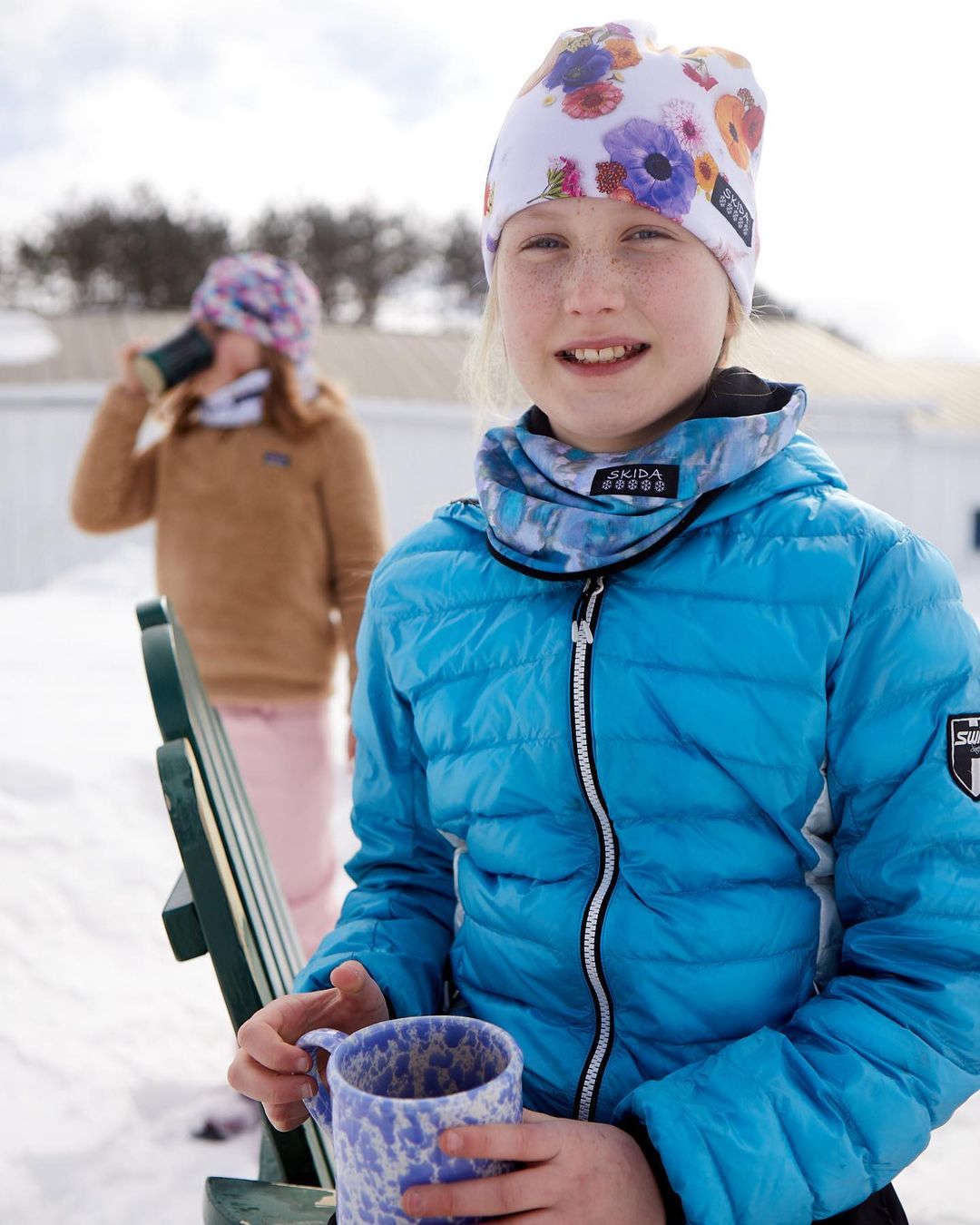 A young girl with a floral beanie and blue puffer jacket is smiling while holding a mug in a snowy, outdoor winter setting. 