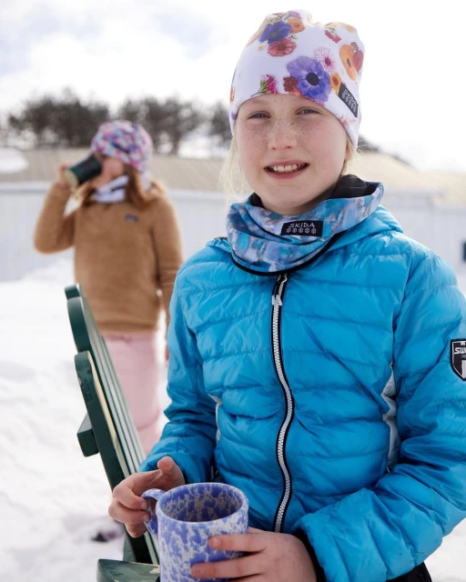 A young girl with a floral beanie and blue puffer jacket is smiling while holding a mug in a snowy, outdoor winter setting. 