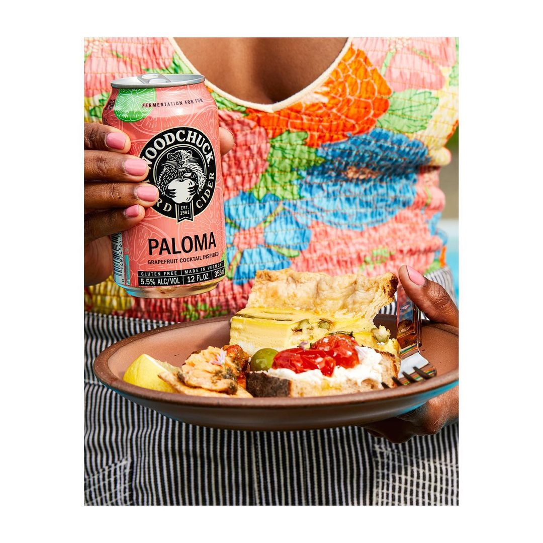 Person holding Woodchuck Paloma hard cider can and a plate with layered savory pie, bread with tomatoes, lemon slice, and an olive.