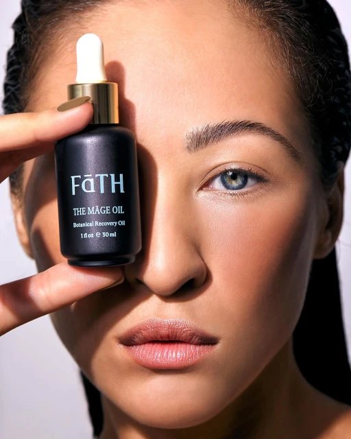 Close-up studio shot featuring an adult woman's face with a black cosmetic oil bottle held by her fingers near her eye.