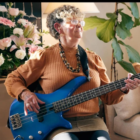 Elderly woman playing a blue bass guitar indoors, wearing an orange sweater and glasses, with flowers and a green plant in the background.