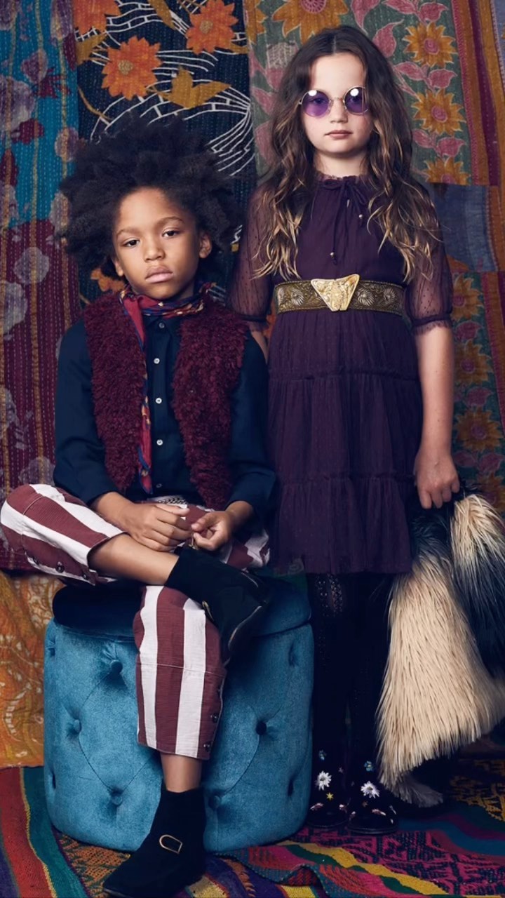 Two children, a boy and a girl, pose in front of a floral quilt. The boy sits on a blue ottoman, and the girl stands next to him.