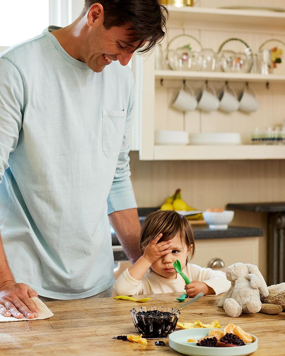 A father and child are at a kitchen counter with fresh fruit and berries. The father is cleaning the counter with a paper towel.