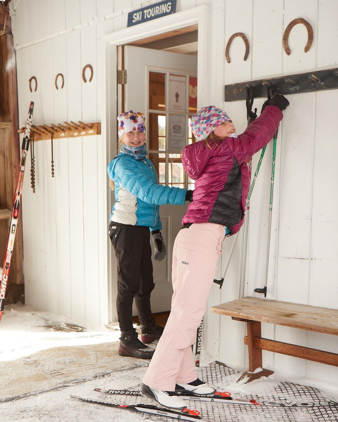 Two young girls preparing for cross-country skiing in winter. One girl in a blue jacket helps the other, who wears a pink jacket, with her poles.